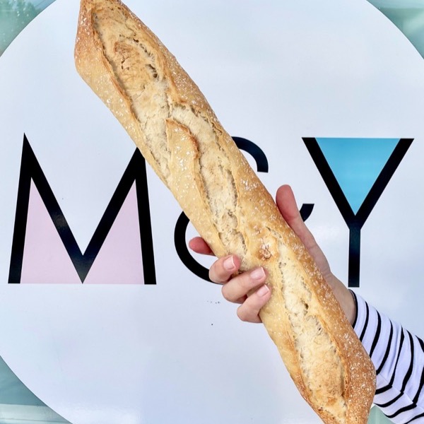A fresh baguette held up in front of the Madame & Yves sign